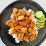 air fryer salmon bites on a black plate on top of white rice next to sliced cucumbers