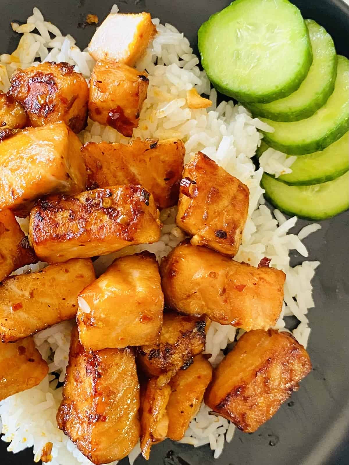air fryer salmon bites on a black plate on top of white rice next to sliced cucumbers
