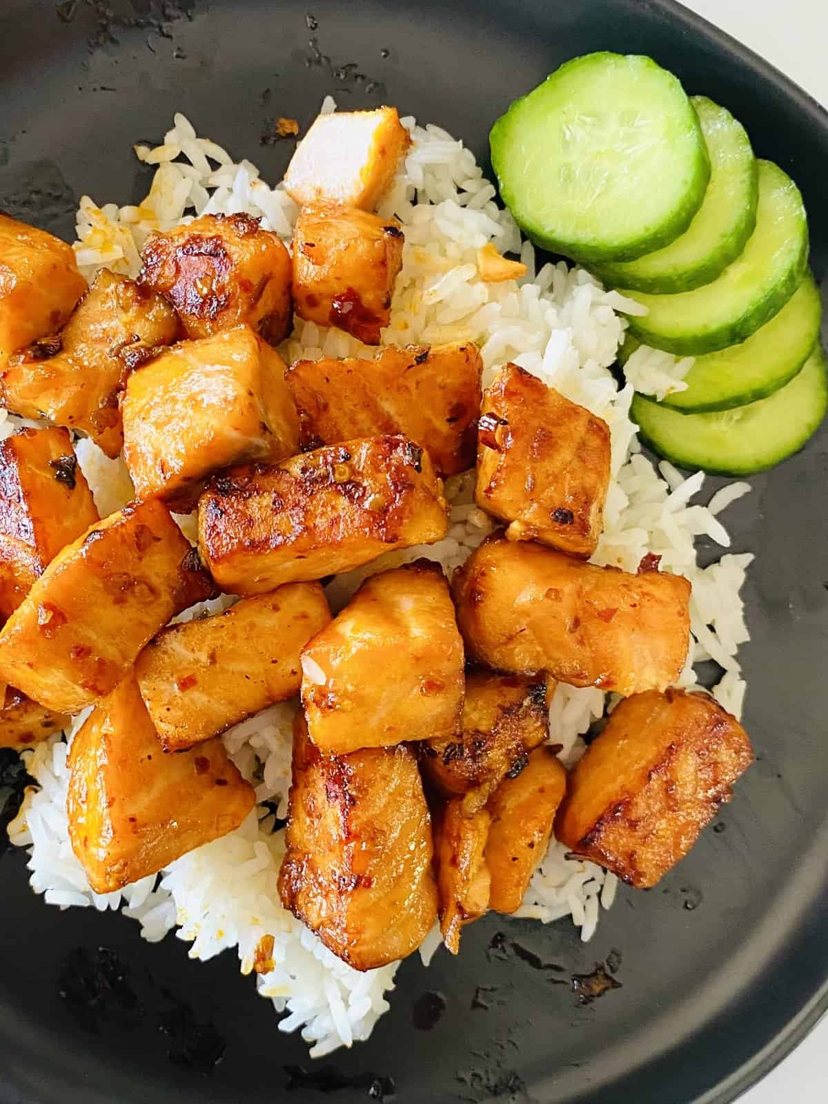 Tik tok air fryer salmon nuggets on a black plate on top of white rice next to sliced cucumbers. 